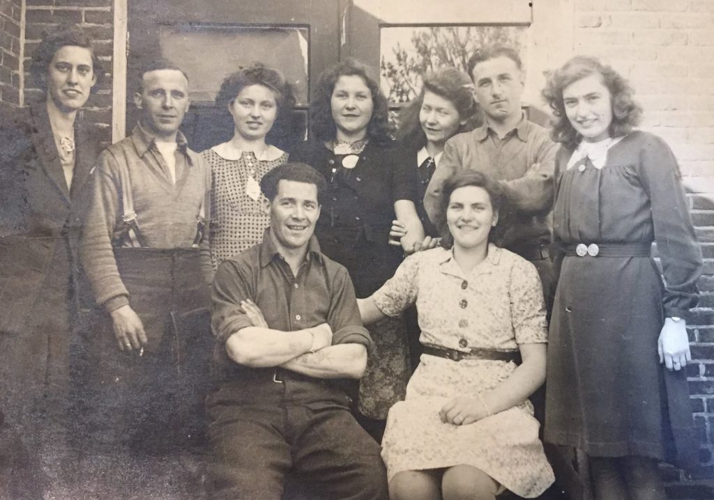A black and white photo. My grandad, Albert, sits front and centre smiling with his arms crossed. He is a white male in army uniform with really cool brylcreemed black hair. Around him stand 8 other people - a mix of young men and women, smiling.